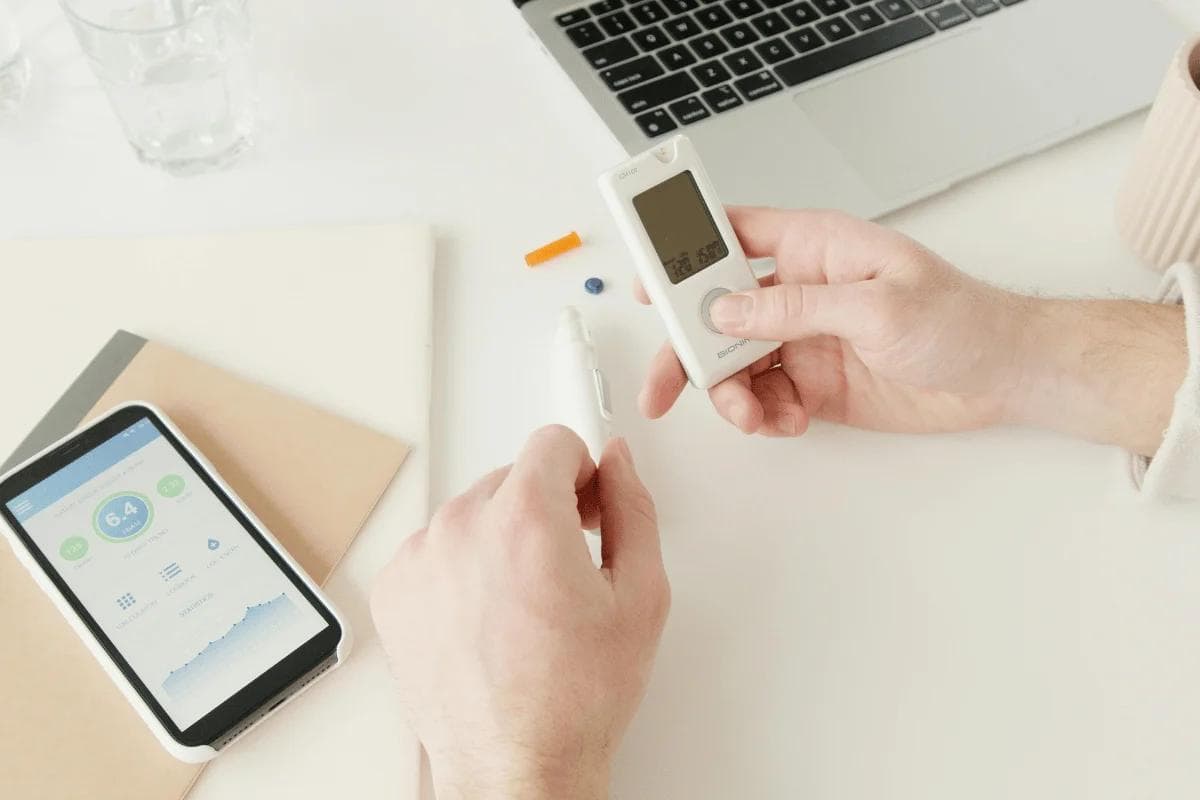 Hands holding a blood glucose meter and lancet, with a smartphone displaying health data, a laptop, and stationery on a white surface.