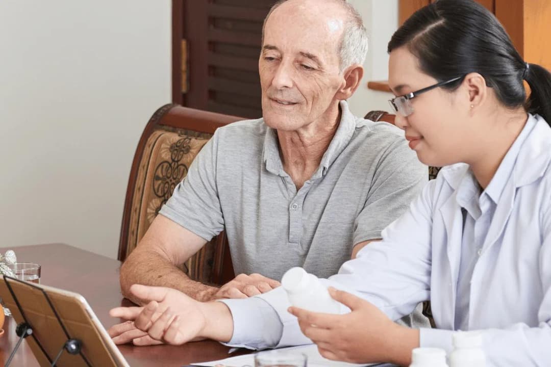 A healthcare professional discusses medication with an elderly man, both focused on a tablet.