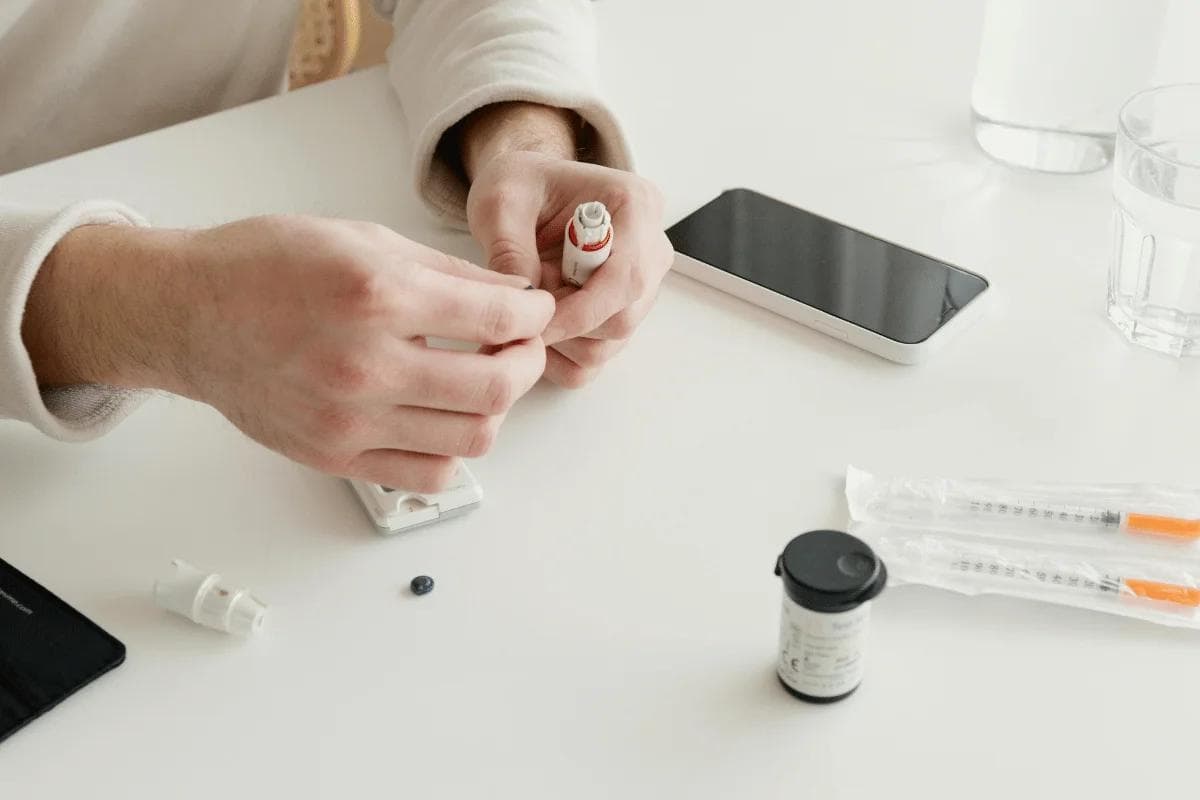 A close-up of a person's hands preparing a medication with syringes and a vial on a table, alongside a smartphone and a glass of water.