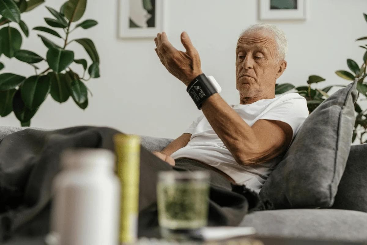 Senior man using digital arm blood pressure cuff while relaxing on sofa.