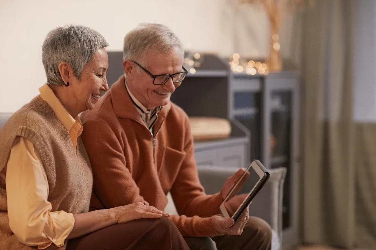 Elderly couple smiling while looking at a tablet together in a cozy living room, creating a warm and joyful atmosphere.